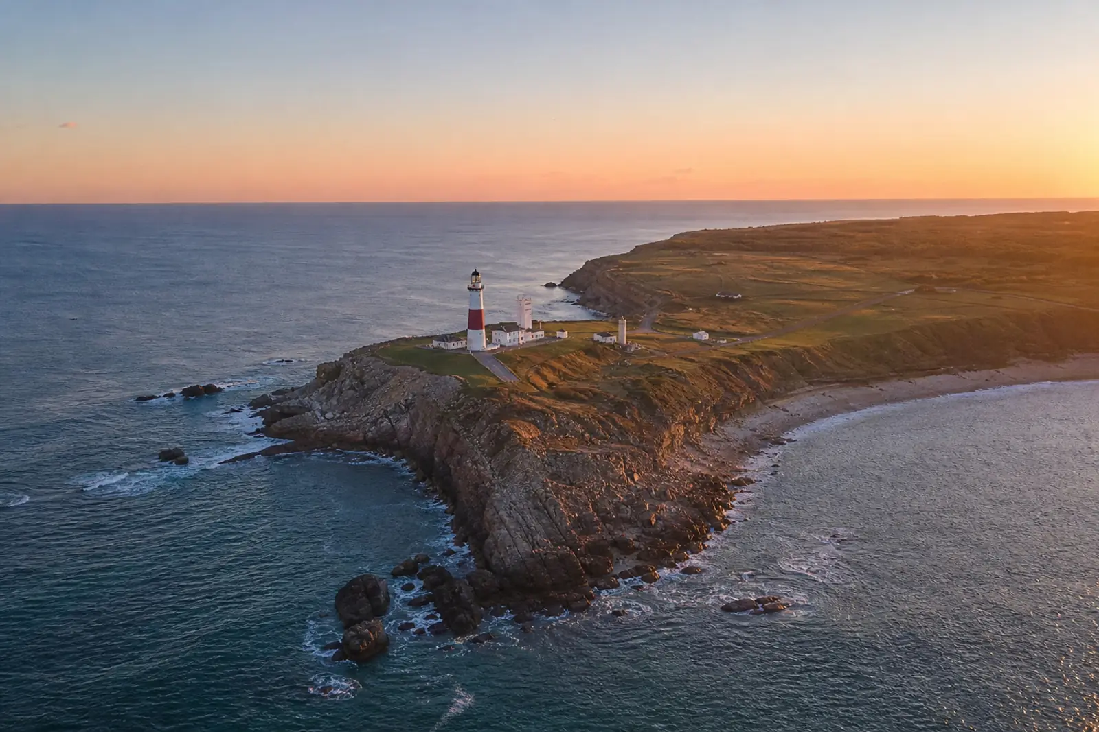 Photo aérienne au couchant d’une pointe rocheuse surmontée d’un phare, prise de haute altitude, où la texture de la roche et les rides de l’eau restent finement détaillées.