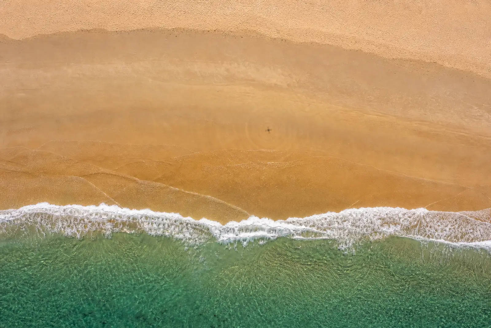 Vue aérienne à la verticale d’une plage déserte au couchant : l’ombre du drone dessine une petite croix sur le sable, au centre des lignes concentriques laissées par les vagues.