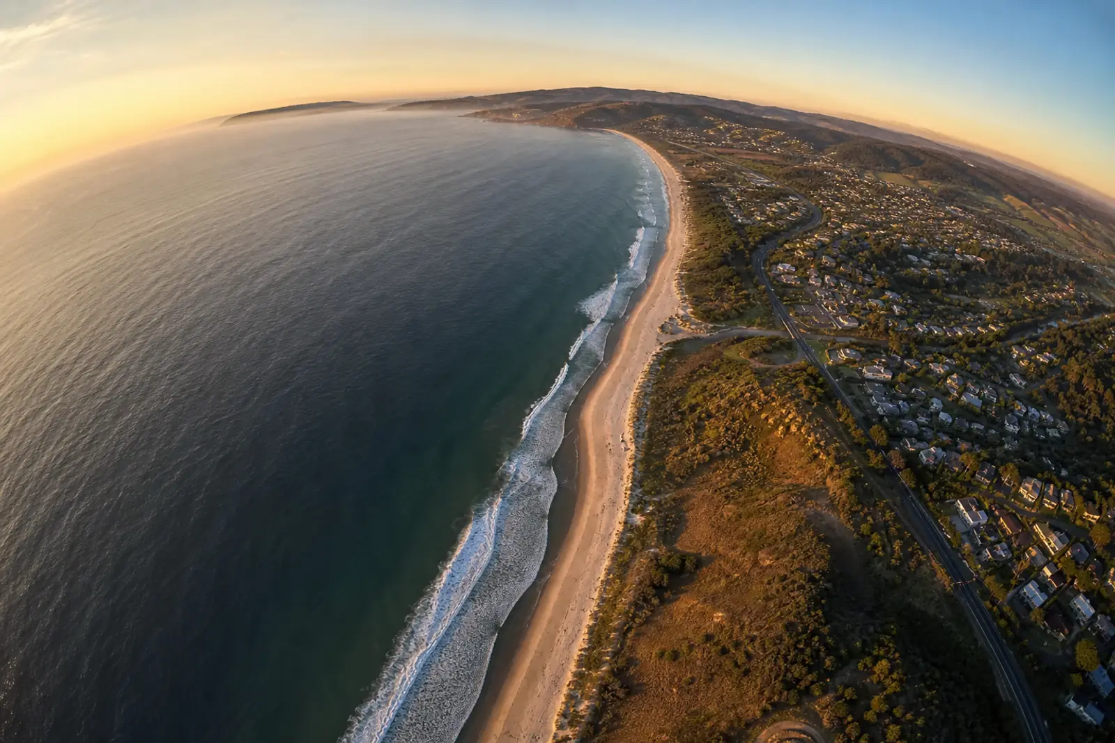 Vue aérienne au couchant d’un long littoral courbe, prise au très grand angle : l’horizon se courbe nettement sur les bords du cadre.