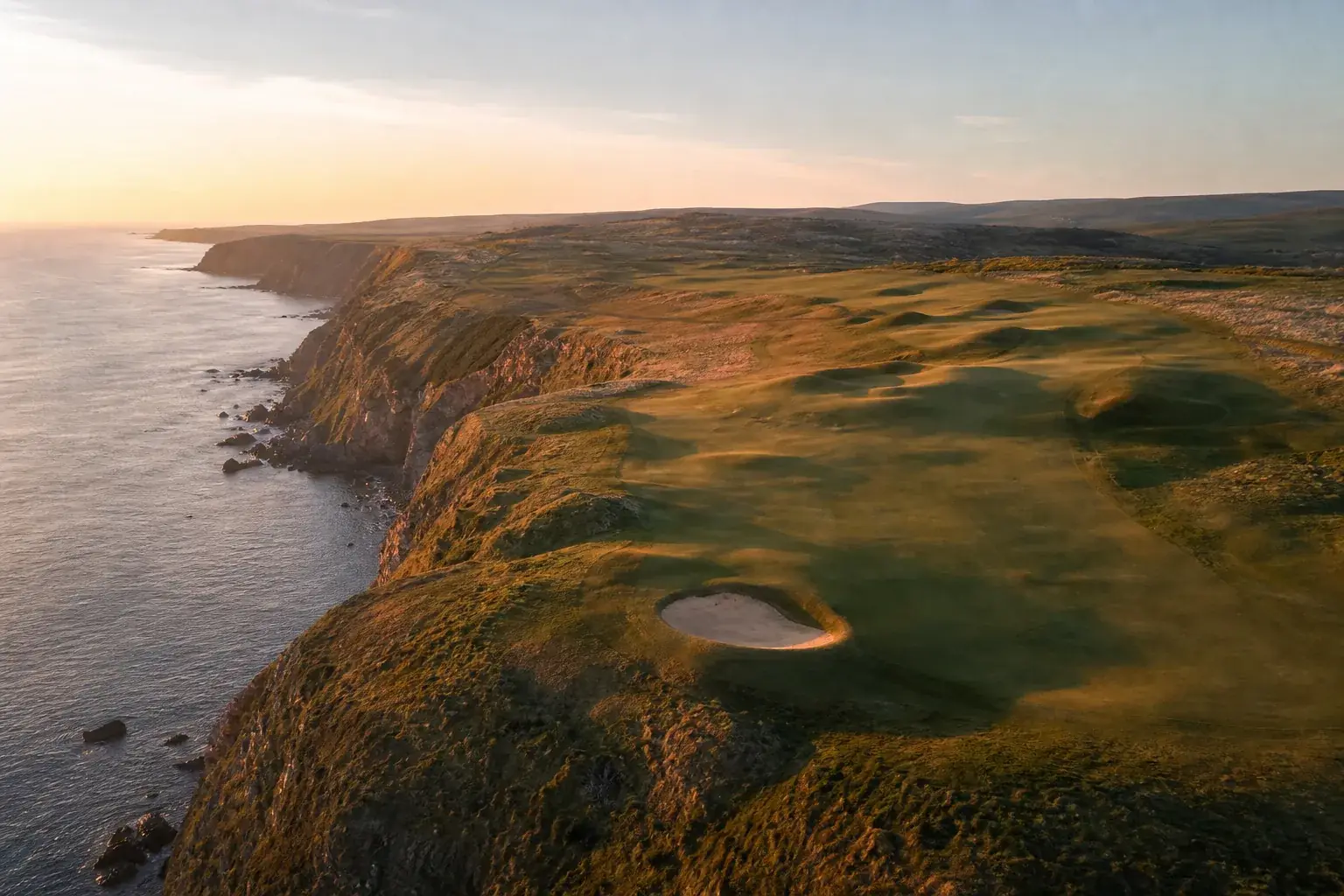 Parcours de golf en bord de falaise au lever du soleil, les fairways suivant le trait de côte, la mer en contrebas.