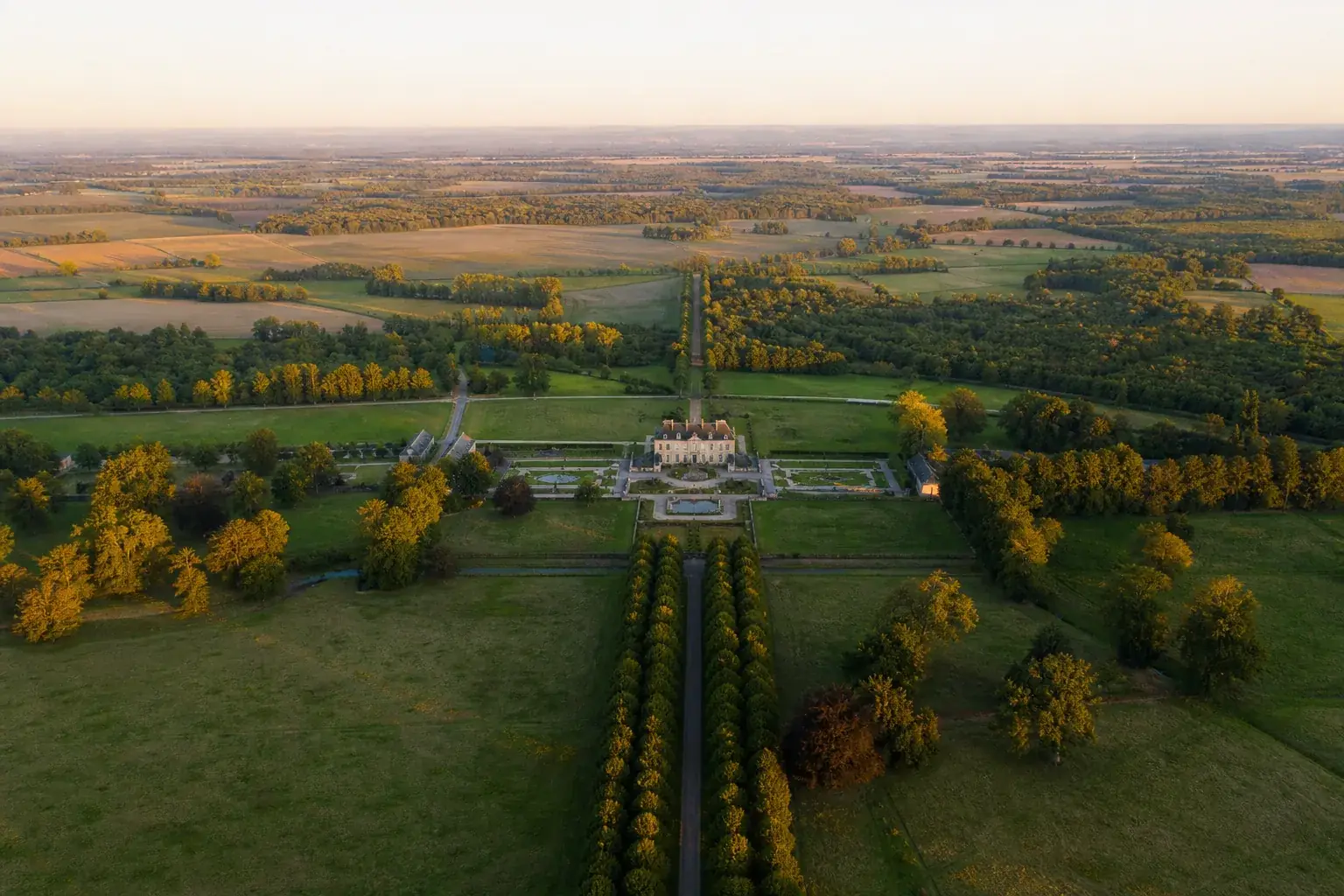Grand domaine de campagne vu du ciel à l’aube : le bâtiment principal au centre, une allée d’arbres et des prairies qui s’étendent bien au-delà.
