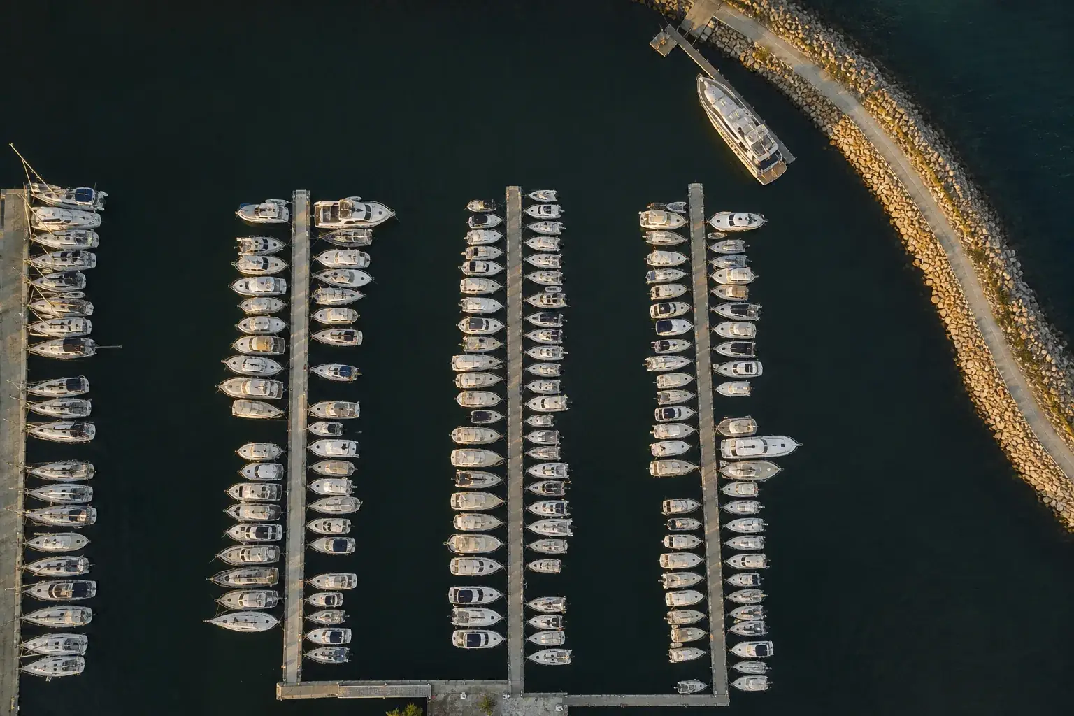 Port de plaisance vu à la verticale : les pannes en lignes parallèles, les coques amarrées, l’eau calme et la digue de pierre.
