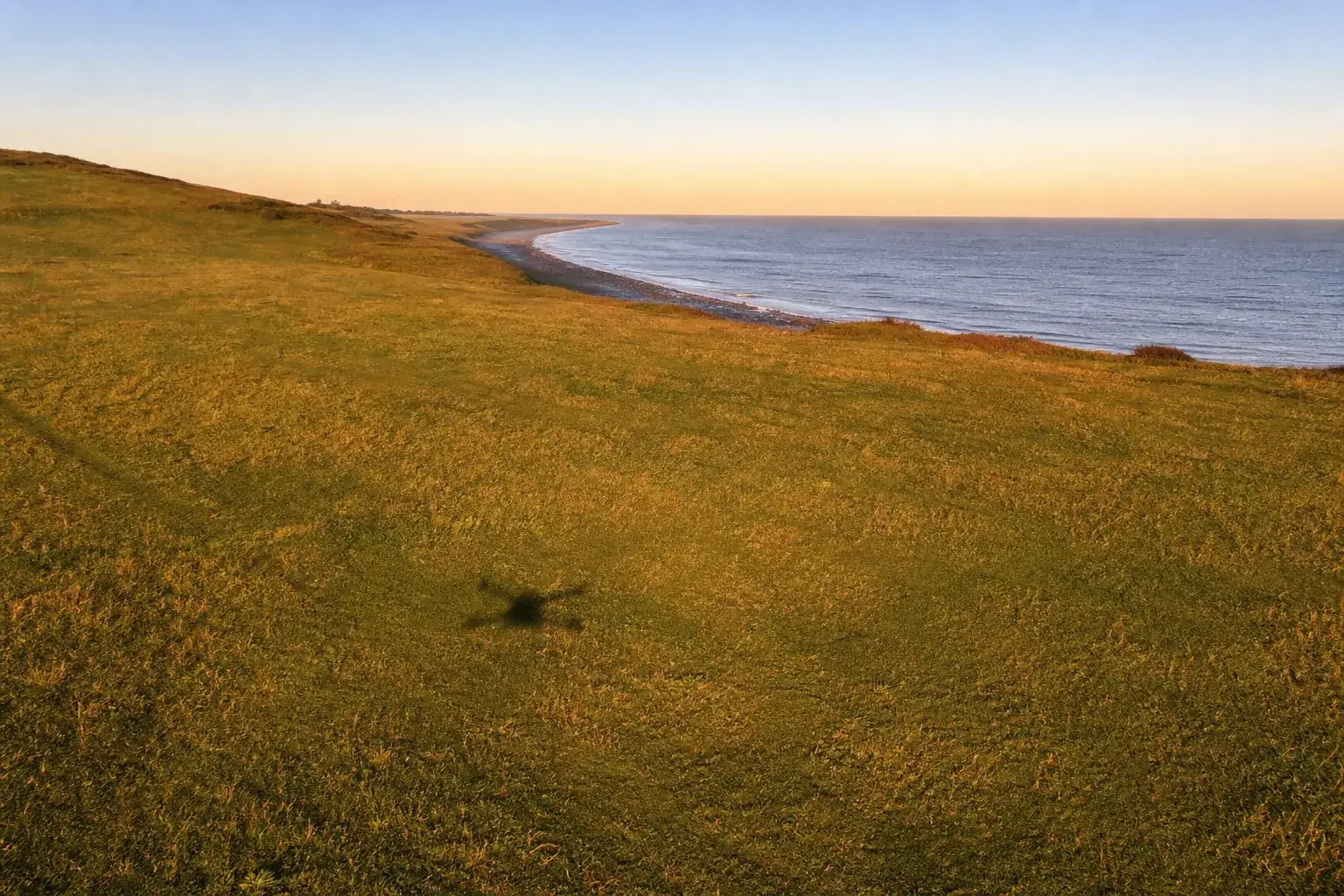 Prairie côtière au lever du soleil, l’ombre d’un petit drone projetée sur l’herbe, le trait de côte qui s’éloigne.