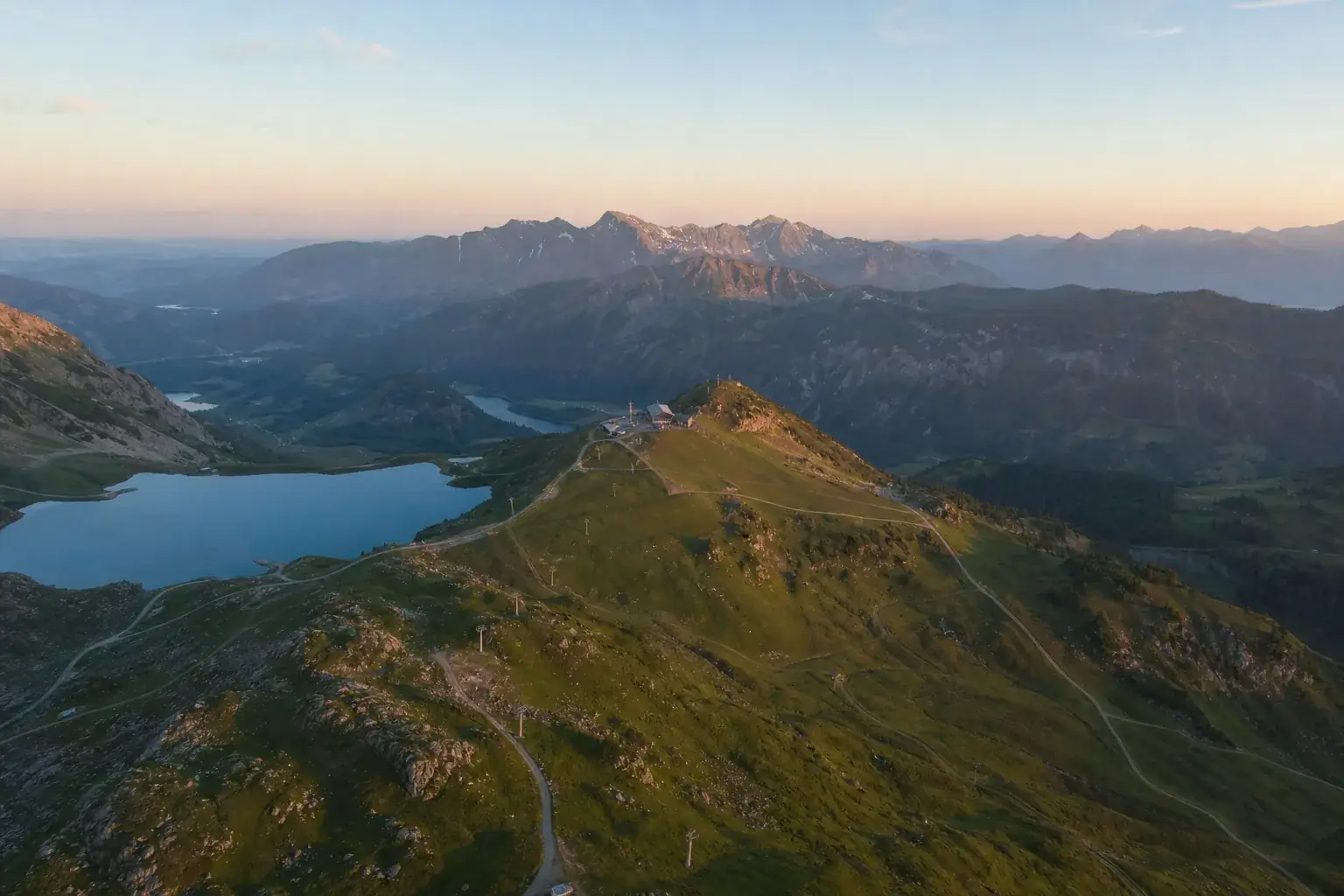 Station de montagne l’été vue du ciel : un lac d’altitude, les pylônes d’une remontée sur les pentes vertes, des sentiers vers une crête.