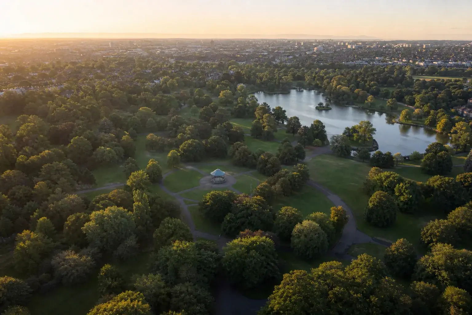 Grand parc public vu du ciel à l’aube : des allées courbes entre pelouses et arbres, un lac d’agrément, un kiosque au centre.