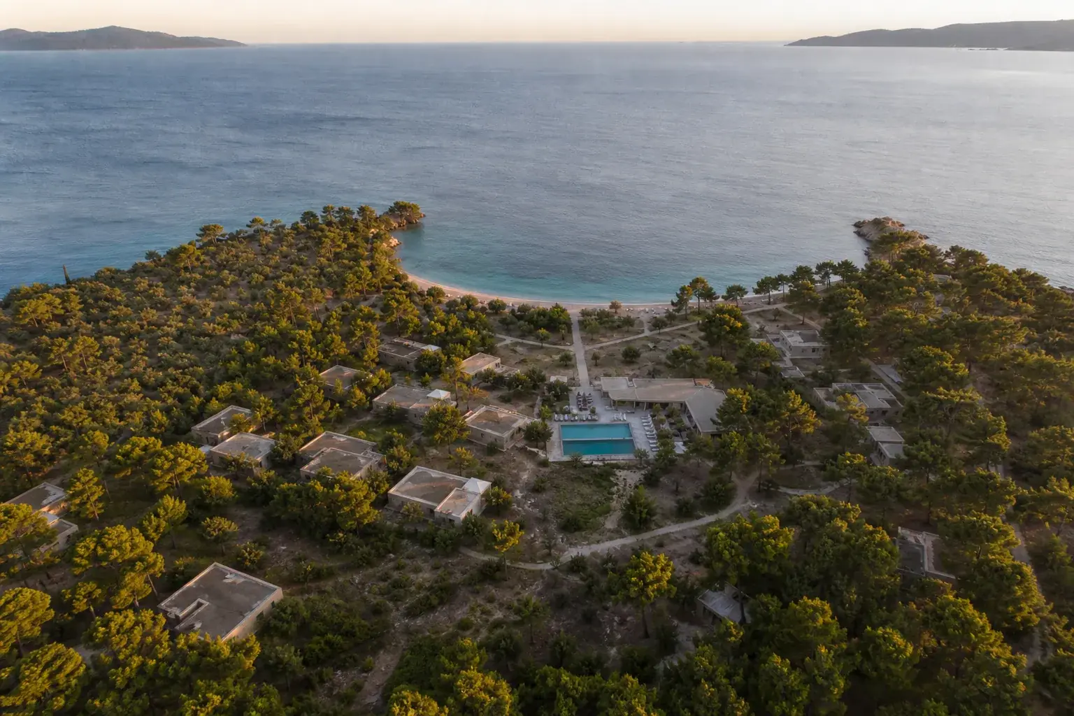 Resort côtier vu de haut : des bâtiments bas dispersés sous les pins, une piscine, un chemin qui descend vers une plage en croissant.