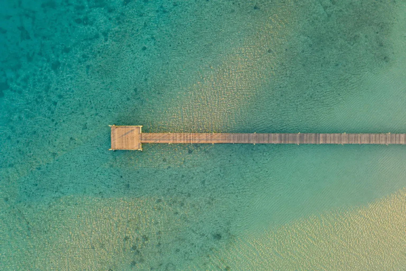 Long ponton de bois s’avançant dans une eau turquoise peu profonde, vu à la verticale, le fond sableux dessiné en dessous.