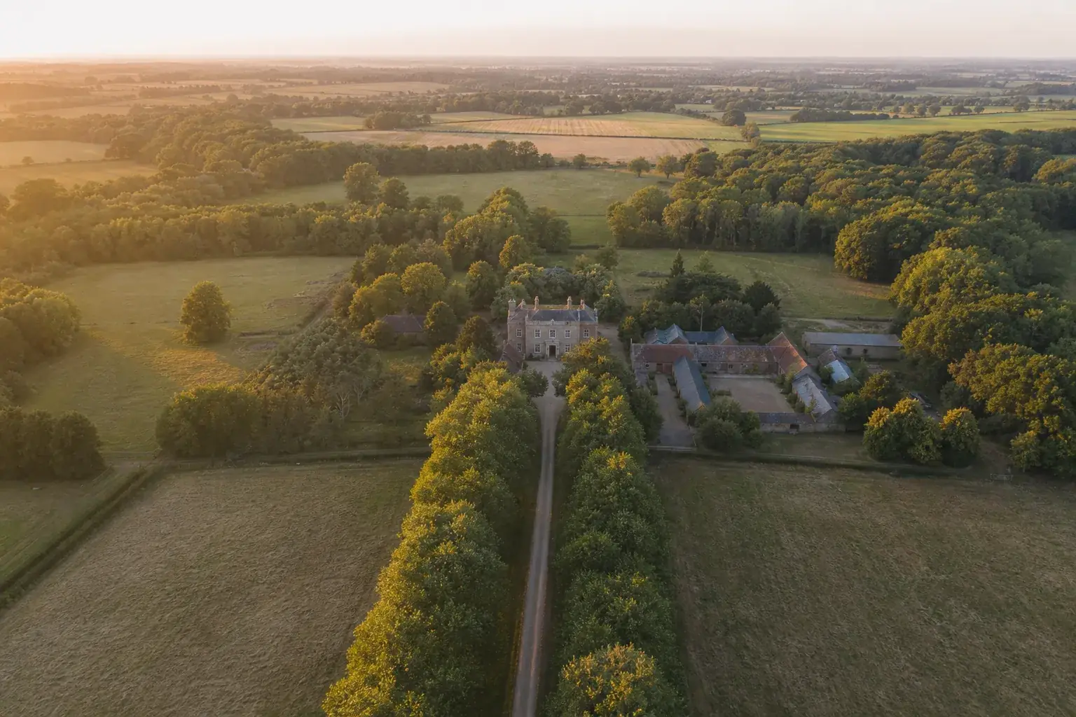 Manoir au bout d’une longue allée bordée d’arbres vu du ciel aux premières lueurs, les dépendances autour d’une cour de gravier.