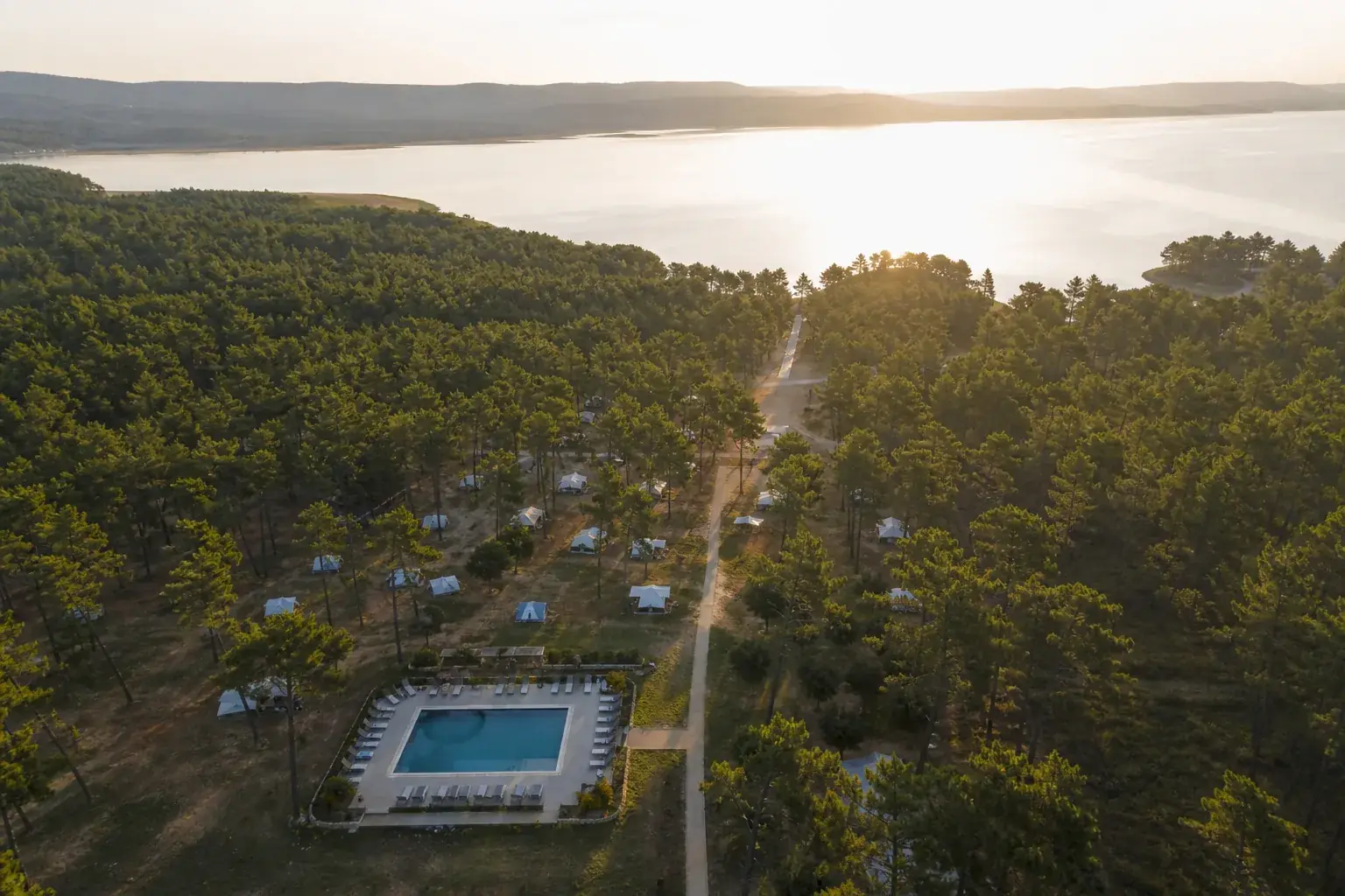 Camping vu du ciel à l’aube, les emplacements dispersés sous les pins, une piscine et un chemin descendant vers un lac.