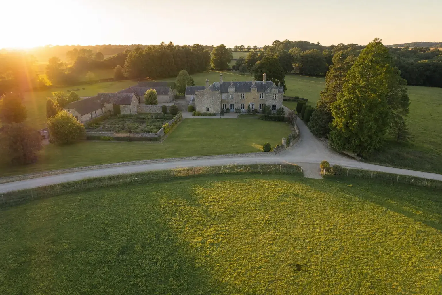 Domaine avec ses bâtiments et ses allées vu du ciel au lever du jour, l’ombre d’un drone posée sur la prairie au premier plan.