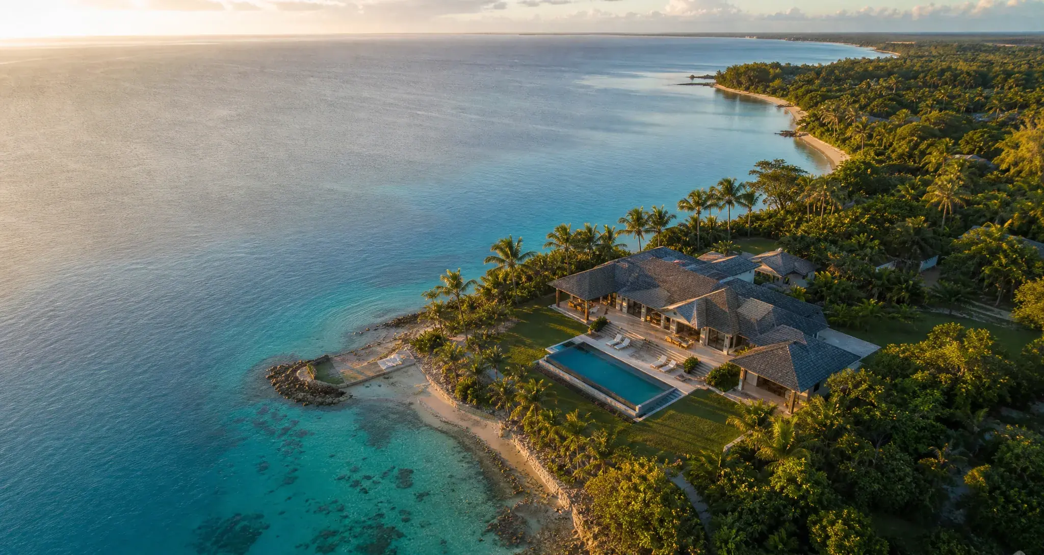 Vue drone d’une villa en bord de lagon turquoise à l’heure dorée, la côte se prolongeant vers l’horizon.