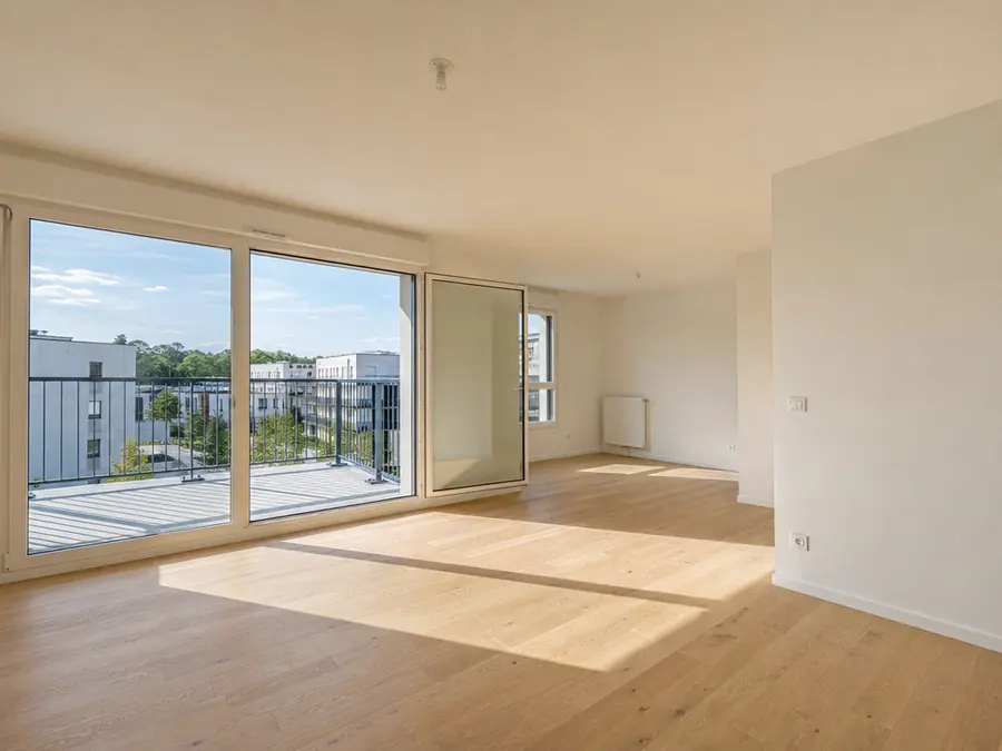 Séjour vide d’un appartement neuf : parquet clair, baie vitrée ouverte sur le balcon et la résidence, lumière du matin sur le sol.