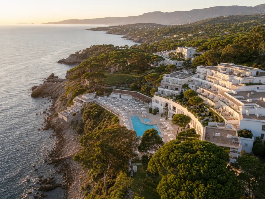 Resort en bord de mer vu du ciel à l’heure dorée : bâtiments blancs en terrasses, piscine et pins parasols au-dessus d’une crique.