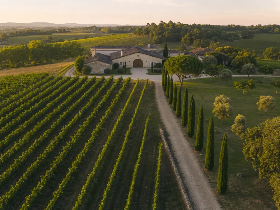 Domaine viticole vu du ciel : rangs de vigne convergeant vers le chai de pierre, alignement de cyprès et collines.